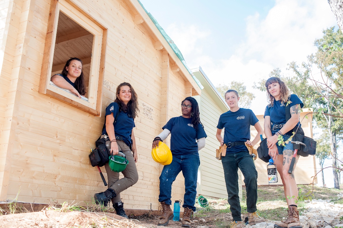 Photo of 5 people at a construction training site, looking at the camera and standing next to a newly built home