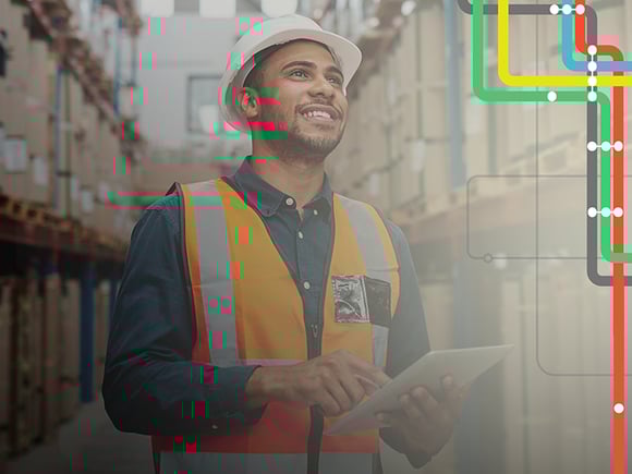 Man wearing a hard hat and safety vest holds a tablet next to shelves of packages
