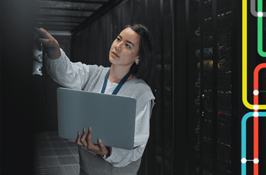 Woman working in a warehouse holds a laptop while pointing to a server on a rack