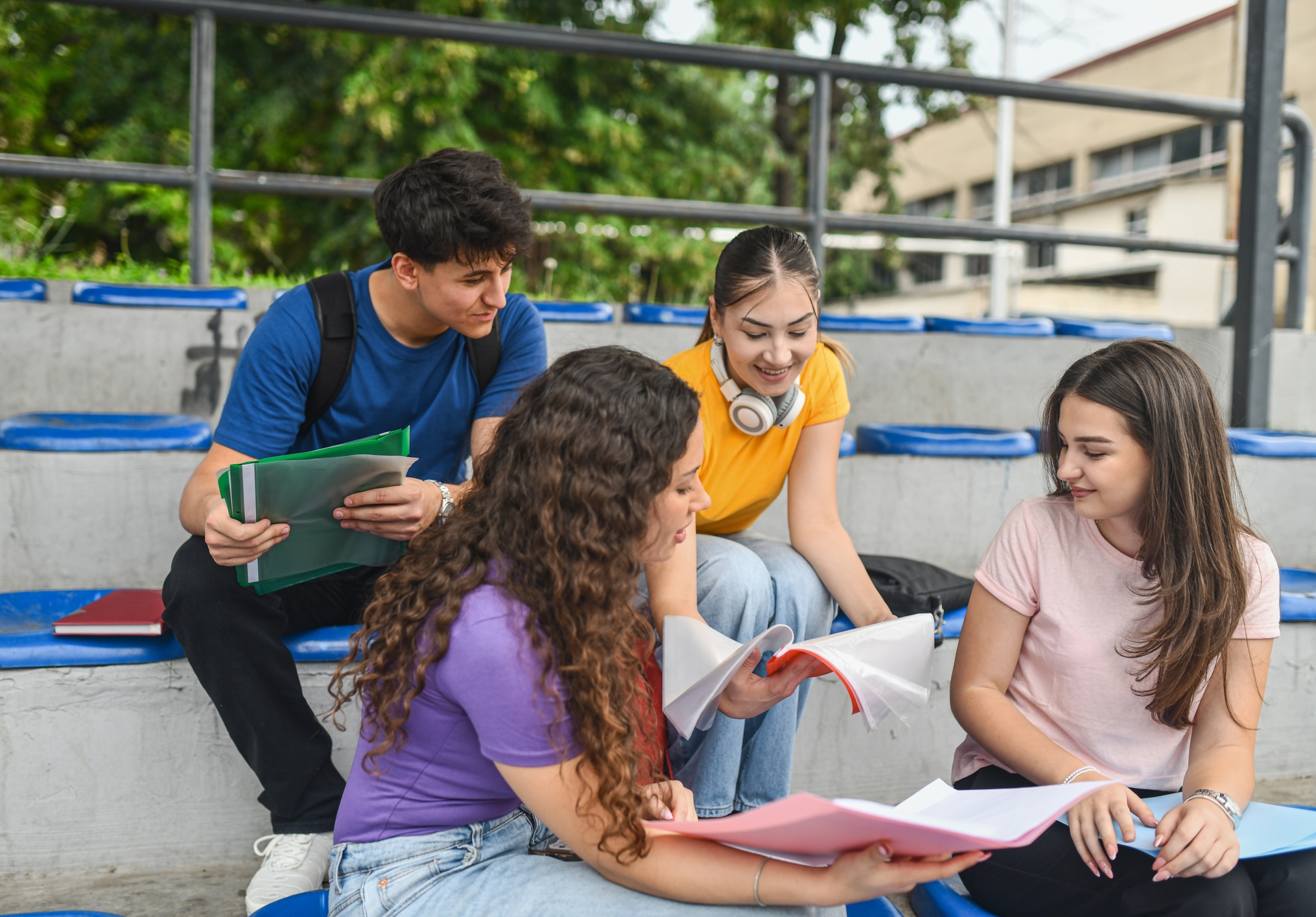 Four students sit on outdoor stadium bleachers looking at open folders and papers.
