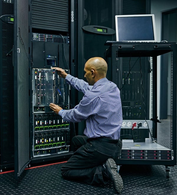 Man kneeling down in a dark server room, replacing wires. 