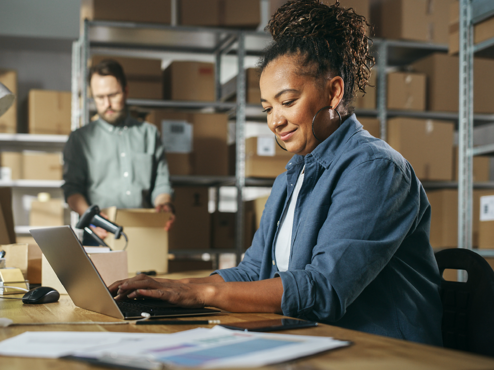 Diverse Male and Female Warehouse Inventory Managers Talking, Using Laptop Computer and Checking Retail Stock. Rows of Shelves Full of Cardboard Box Packages in the Background.