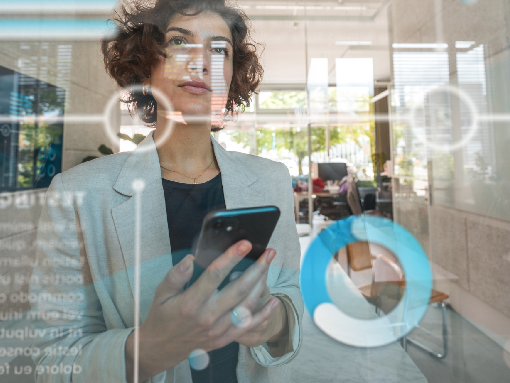 Caucasian businesswoman standing in front of a transparent screen in a modern office. Analyzing business data on screen using a smartphone.